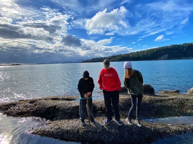 Three children stand on a barnacle covered rock looking down at tide pools.