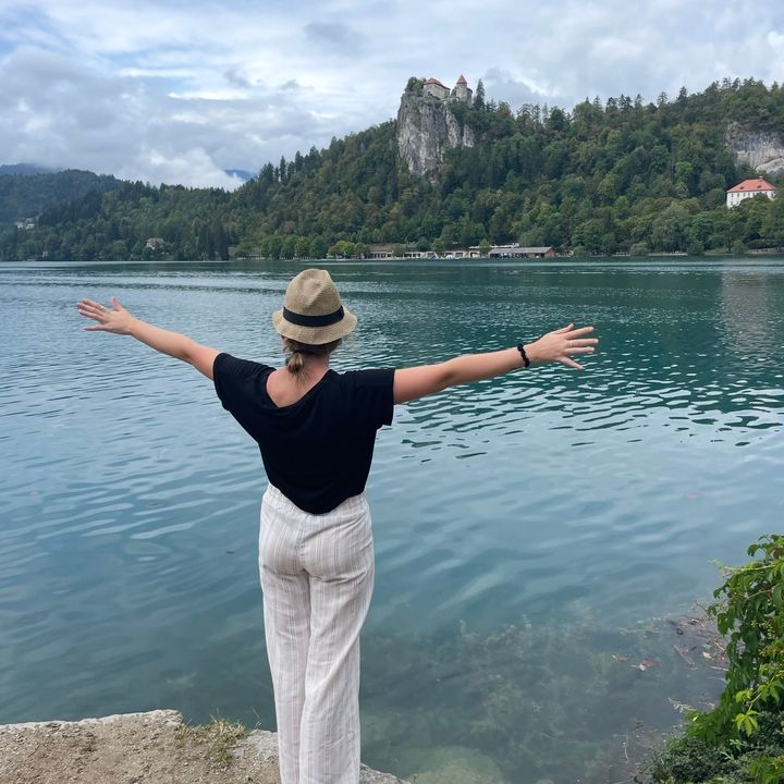 Blond caucasian woman wearing a bucket hat looks out over a blue lake with green trees and a castle perched in the hills behind it.
