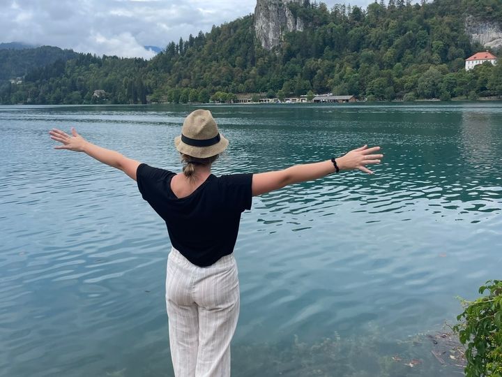 Blond caucasian woman wearing a bucket hat looks out over a blue lake with green trees and a castle perched in the hills behind it.