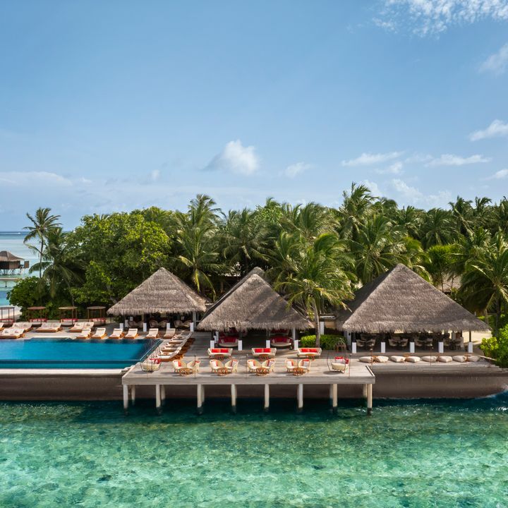 A resort area with thatched roofs and an infinity pool surrounded by palm trees on a platform over the water at Anantara Veli Maldives Resort.