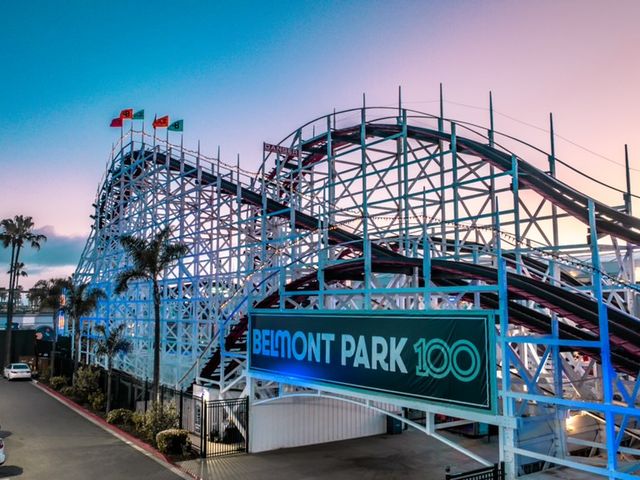  A rollercoaster sits behind a Belmont 100 banner with palm trees in the foreground and background with a pink dusk sky.