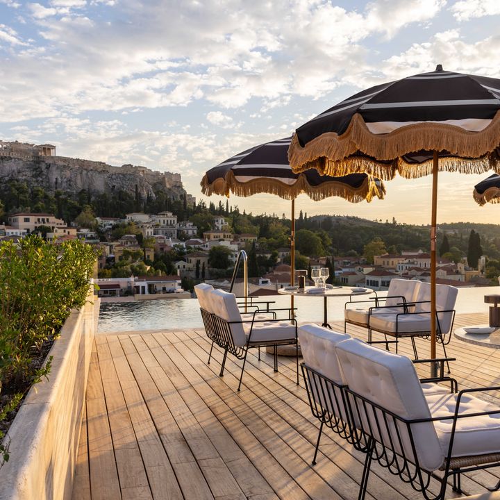 Outdoor dining tables with fringe-trimmed umbrellas on a wood deck overlooking water and ancient buildings in Athens, Greece at THE DOLLI at Acropolis rooftop