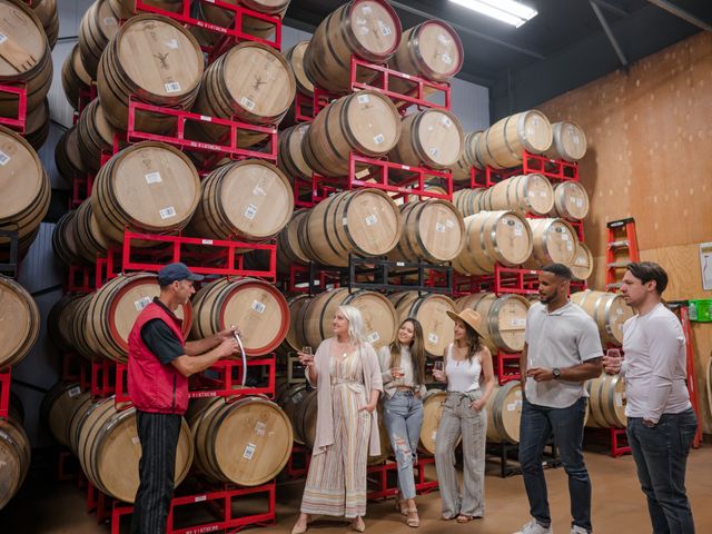 A group of three women and two men listen to a guide on a winery tour next to floor to ceiling red racks holding wine barrels.
