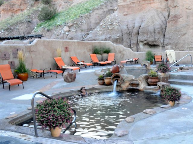 A person soaks in a mineral hot springs bath amid a Southwestern landscape with orange pool loungers surrounding the bath