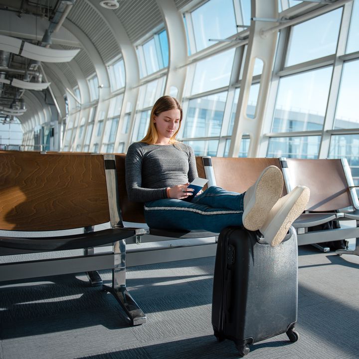 Blonde woman with documents and luggage sitting in the empty airport terminal hall while waiting her flight.