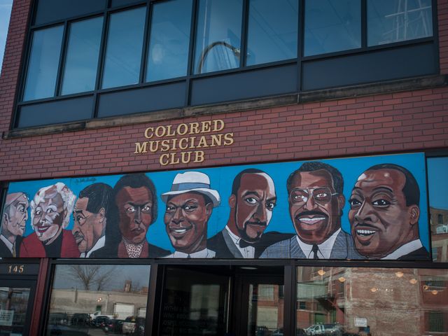 Daytime view of the brown brick exterior of the Colored Musicians Club in Buffalo. A mural of notable Black musicians from its history hangs above the doorway.