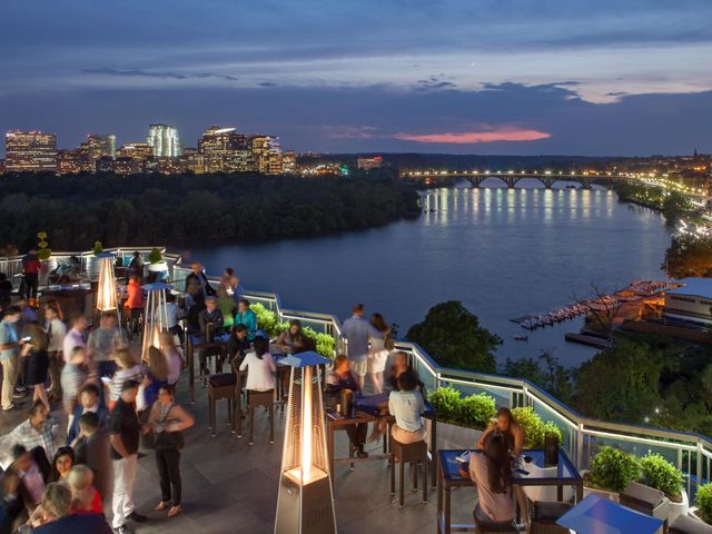 A bustling crowd at the Top of the Gate rooftop bar atop the Watergate Hotel at nighttime with skyline views of Washington D.C.