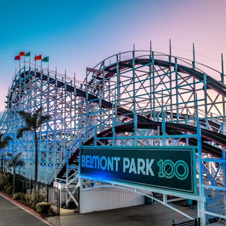 A rollercoaster sits behind a Belmont 100 banner with palm trees in the foreground and background with a pink dusk sky.