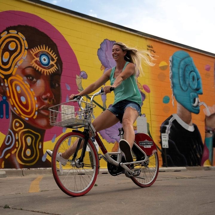A woman rides a bike next to a colorful mural covering the entire exterior wall of a building.