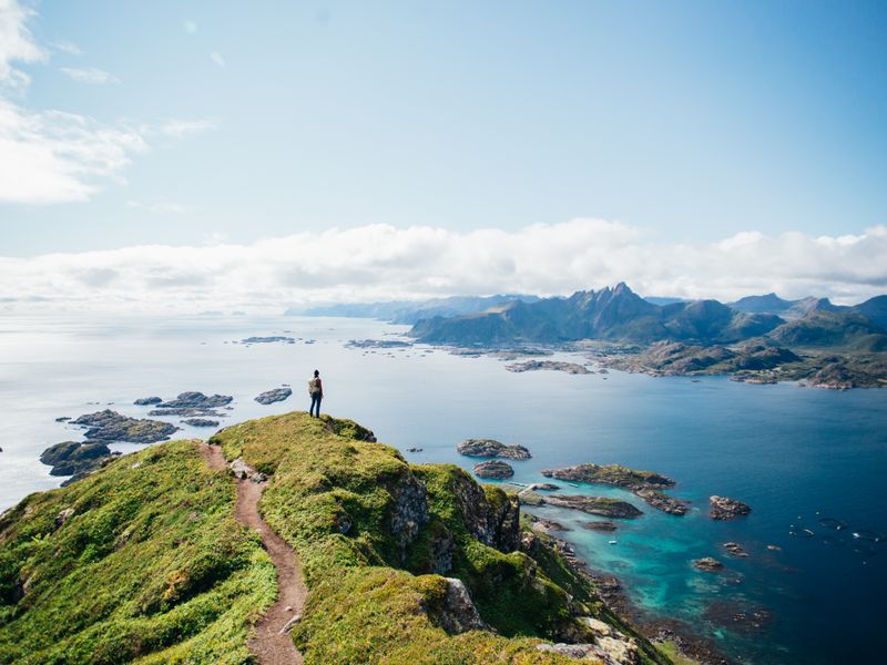 A hiker stands on top of a mountain after a hike in Lofoten Islands in Norway looking out over rocks, water, and mountains.
