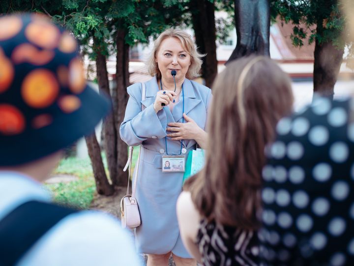 A female tour guide wearing a name tag speaks into a microphone while addressing a group.