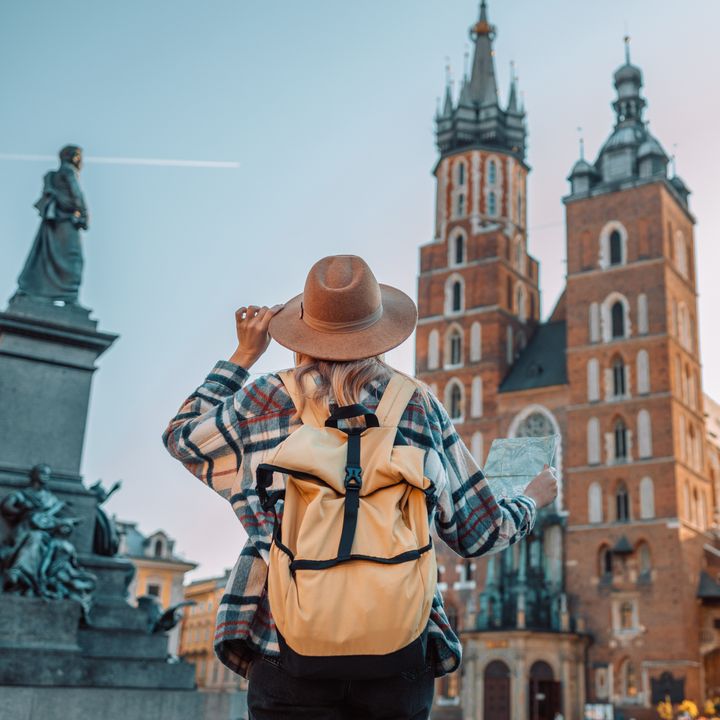 A tourist in Europe wearing a hat and a backpack as seen from behind next to historic buildings.