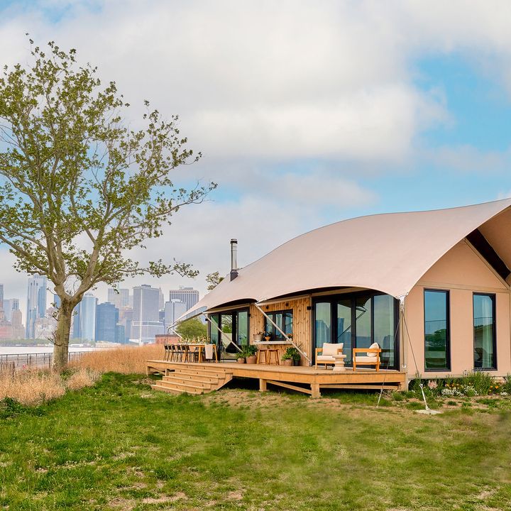 A glamping tent on a grassy area next to a tree with views of the New York City skyline behind it.