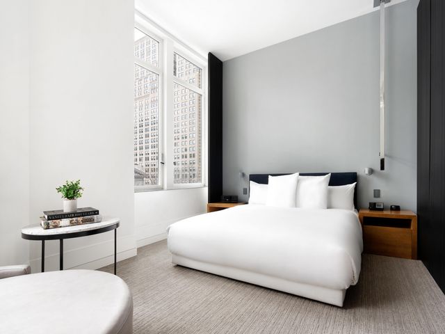 A starkly designed guest room at Andaz 5th Avenue in New York with a white bed, white seating, one white wall, and contrasting grey and black walls