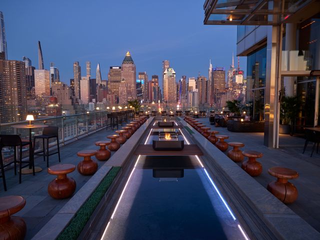 A reflective pool with firepits and seating around it on a NYC rooftop with eastern views of the New York skyline at night.