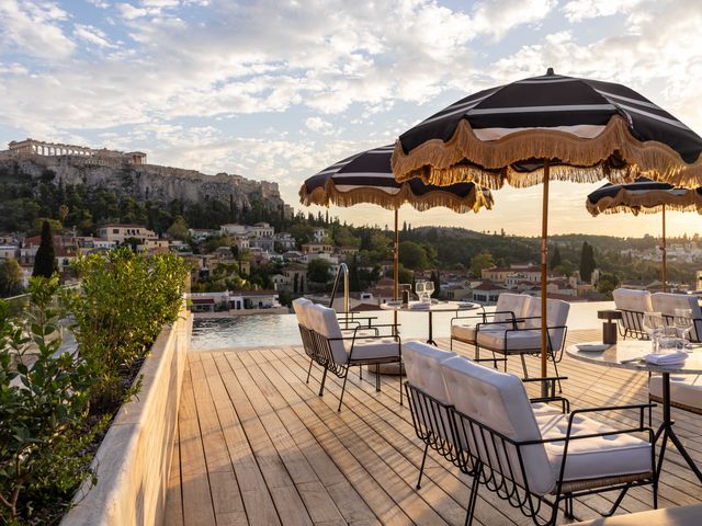 Outdoor dining tables with fringe-trimmed umbrellas on a wood deck overlooking water and ancient buildings in Athens, Greece at THE DOLLI at Acropolis rooftop