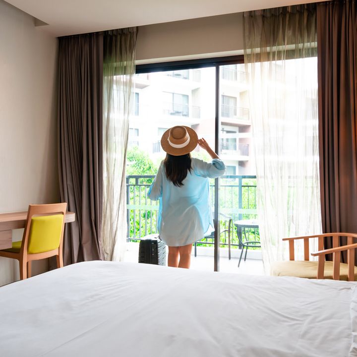 A woman looking out the window of a hotel room wearing a floppy hat and holding her suitcase next to the bed.