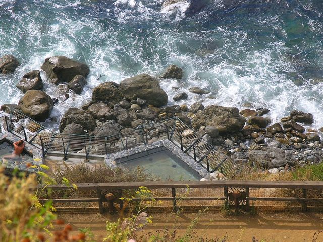 An aerial view of hot springs along rugged oceanside with waves crashing on rock boulders