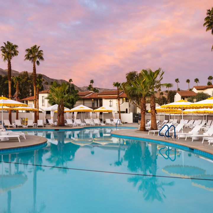 A pool surrounded with white chairs, yellow striped umbrellas, and palm trees at sunset at Omni Rancho Las Palmas Resort & Spa.