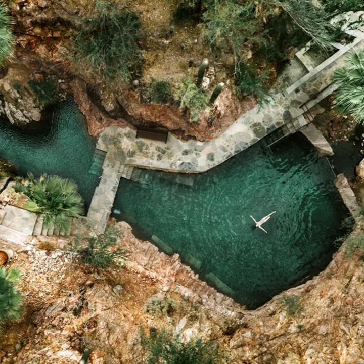 An overhead drone shot of someone swimming in a hot spring surrounded by rocky cliffs and palm trees at Castle Hot Springs.