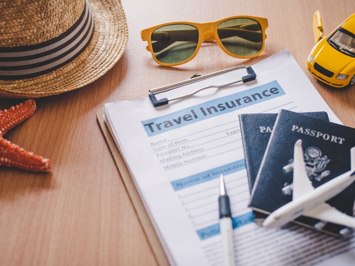 A clipboard with travel insurance paperwork plus passports, a pen, sunglasses, a hat, starfish, and toy plane and car.