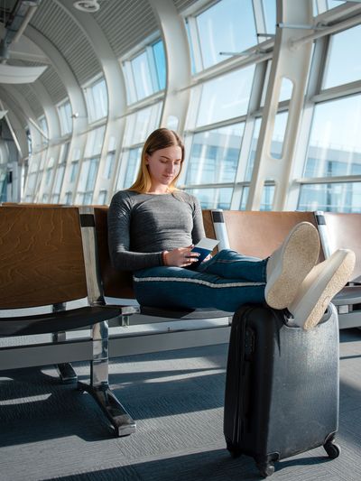 Blonde woman with documents and luggage sitting in the empty airport terminal hall while waiting her flight.