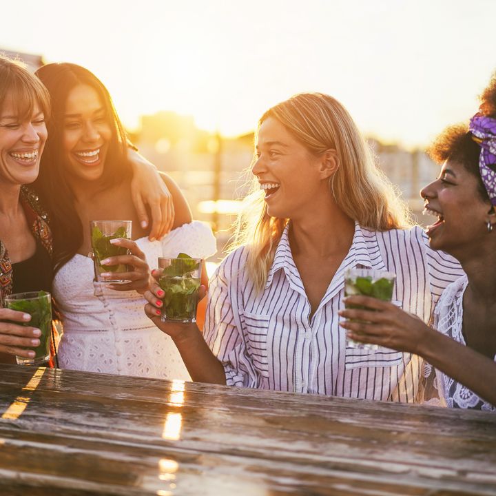 A group of women sitting outside together laughing while all holding drinks.