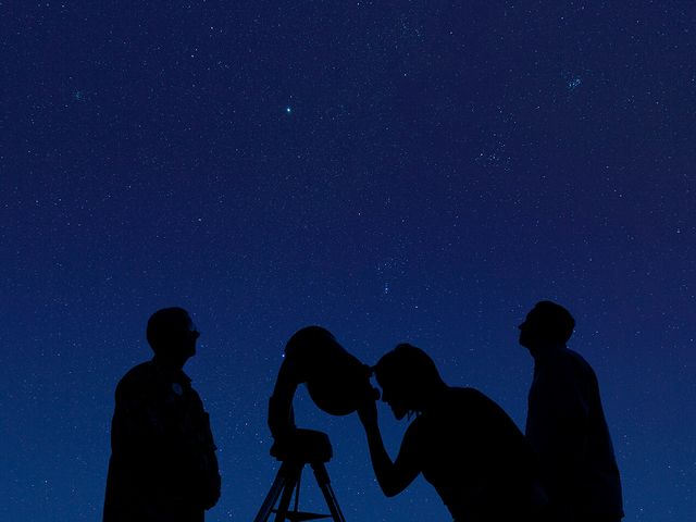 The outline of three people are seen looking into a telescope at night.