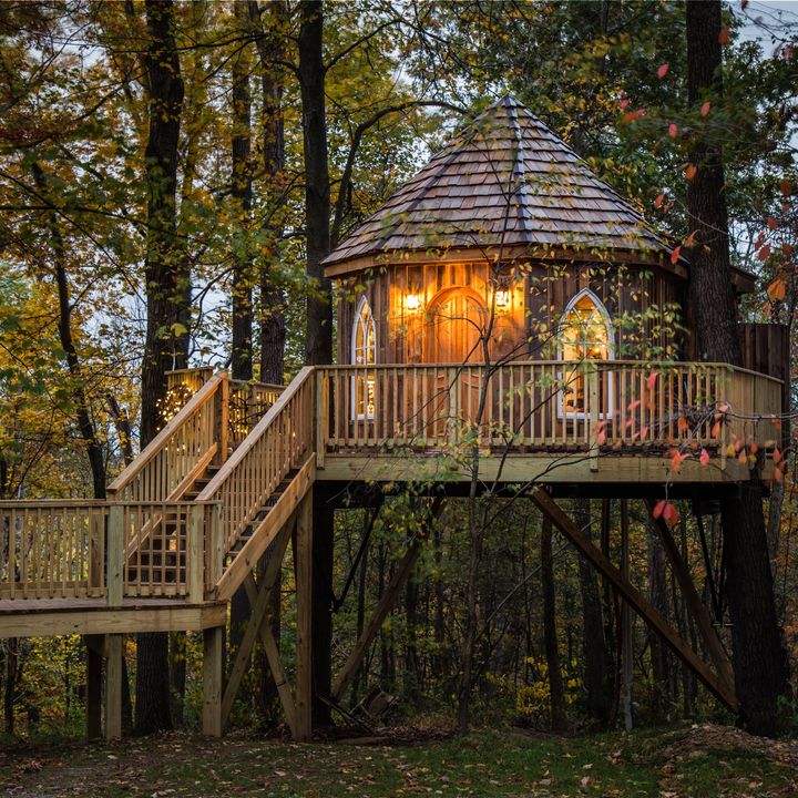 A cylindrical-style treehouse with a shingled roof and stairs leading up to it is perched in the woods.