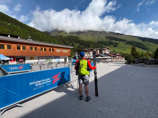 A man standing at the base of Hintertux Glacier in summer holding his skis with green mountains around.