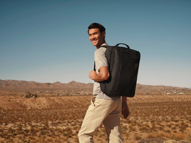 A man smiles for the camera while wearing a large black backpack in a desert setting.