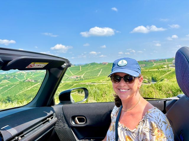 A woman wearing a blue baseball hat sits in a convertible car and smiles at the camera next to scenic vineyards.