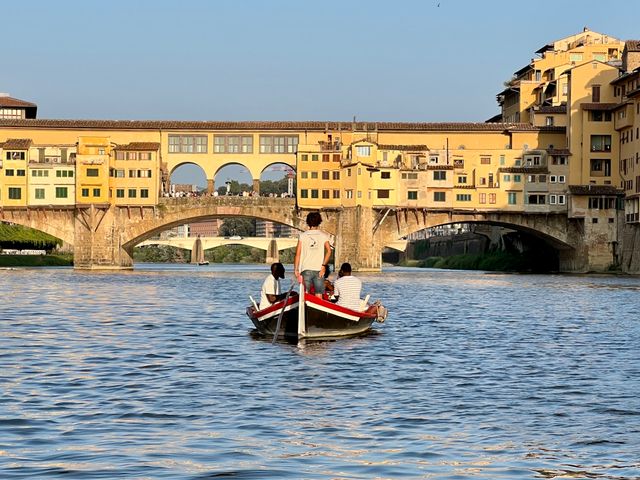 A gondola-style boat on a river passing under the Ponte Vechio Bridge in Italy.