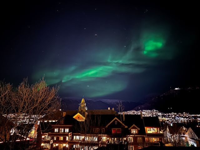 Green northern lights seen in a night sky over the city of Tromso with lights with buildings.