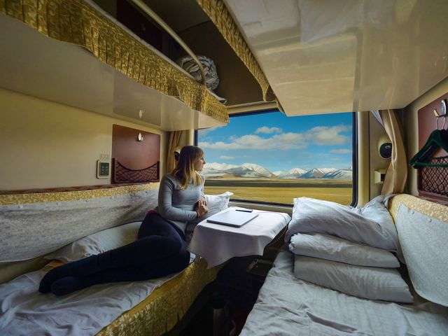 A woman sitting inside a sleeper train car with a mountainout scene in the distance.