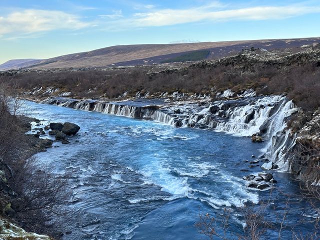 Low waterfalls stream into a river next to bare trees and landscapes dotted with a little snow.