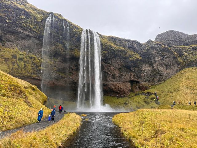 A waterfall cascades down the side of a mountain covered in mossy grass with a walking path behind it where people are visible.