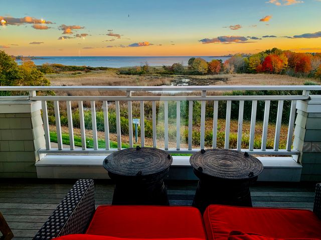 A wooden deck with red cushioned seating facing a marsh and ocean with fall trees in between.