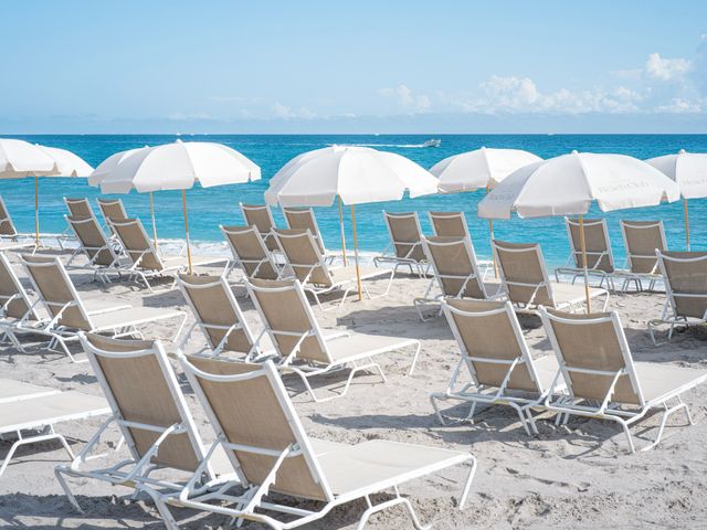 Dozens of white beach chairs with white umbrellas on a white sand beach facing the blue ocean water.