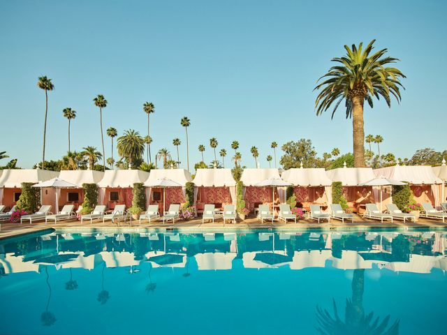 The Beverly Hills Hotel pool with a row of pink cabanas on one side underneath a blue sky and palm trees,.