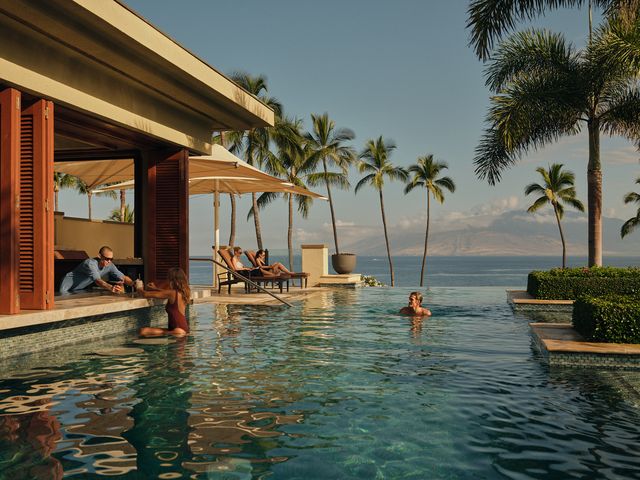 The adults-only infinity pool at the Four Seasons Resort Maui with a few swimmers in the water, someone at the swim-up bar, people on lounge chairs, with ocean and mountain views.