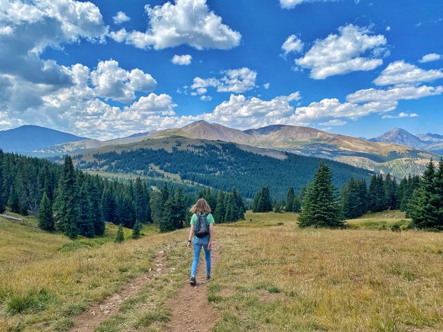 A teenager as seen from behind walking on a mountain trail in front of greenery and peaks.