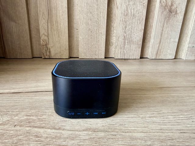 A small black white noise machine with blue trim and blue buttons in front of a wood wall on wood floors.