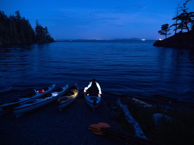 Three empty kayaks sit on the shore of a beach at night next to another kayak with a person inside that is glowing in front along the water.