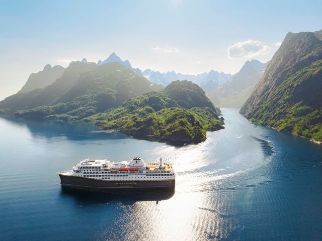 An aerial view over a Havila Voyages ship sailing through mountainous terrain on a sunny day.
