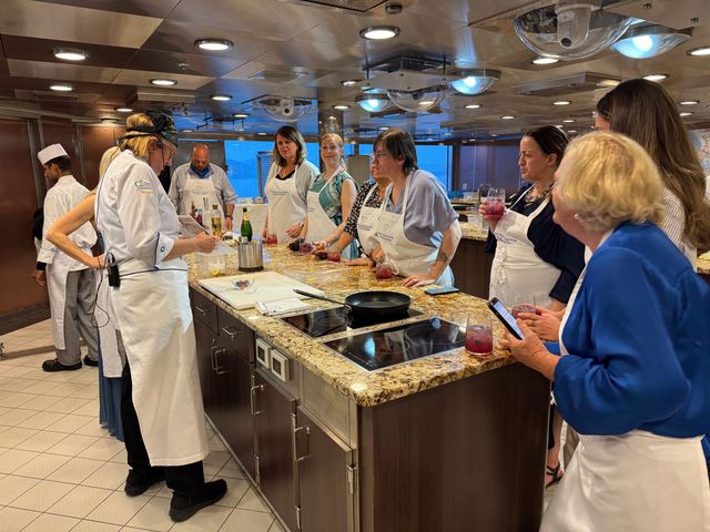 A group of people cluster around a kitchen island with chefs in a cooking class.