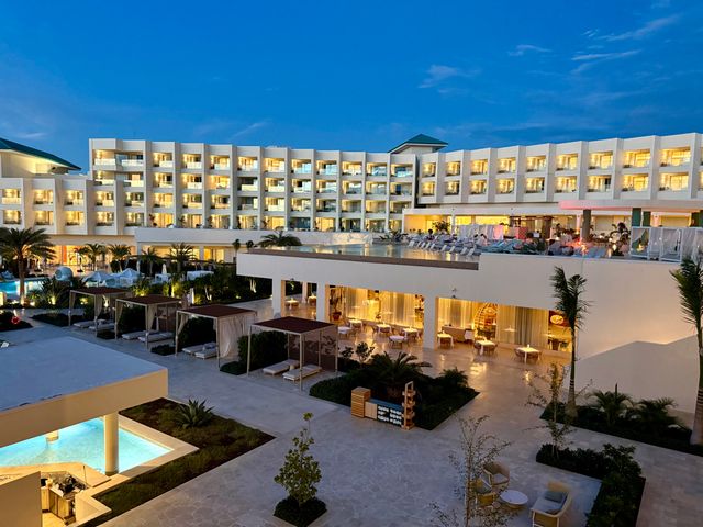 The exterior of the Joia Aruba Iberostar with rows of white balconies, a terrace pool, and a restaurant underneath photographed at dusk.