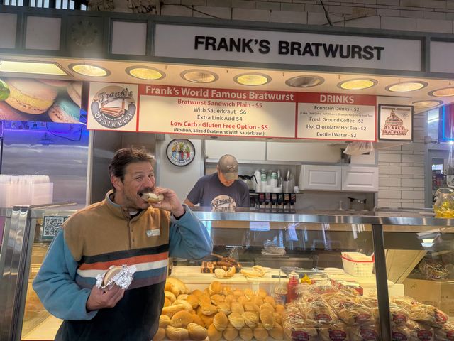 A man bites into food in front of a bratwurst stand. Caption: The author at West Side Market