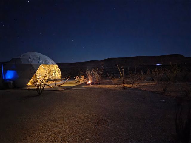 A geodesic dome lodging seen at night with light inside, illuminating a small deck with a hammock and seating on a desert field.