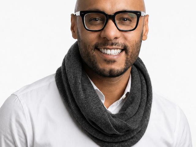 A man wearing glasses and a scarf around his neck, smiling for the camera against a white background.